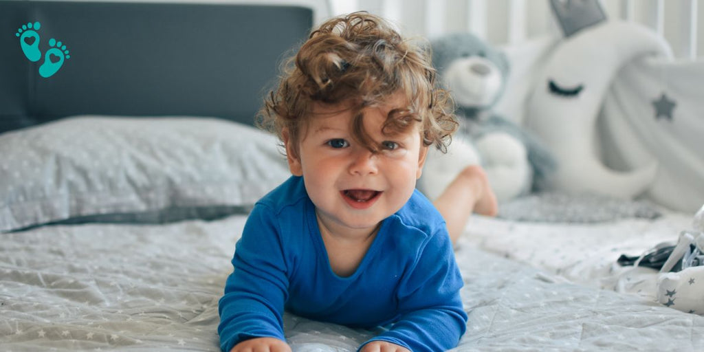 Curly-haired baby in a blue onesie smiling on a bed with plush toys, perfect for Grookz baby shoes