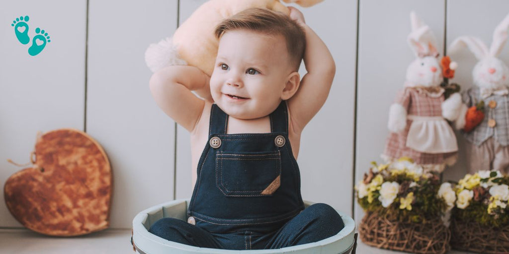 Smiling baby in denim overalls sitting in a basket with toys, showcasing the comfort and style of Grookz baby shoes