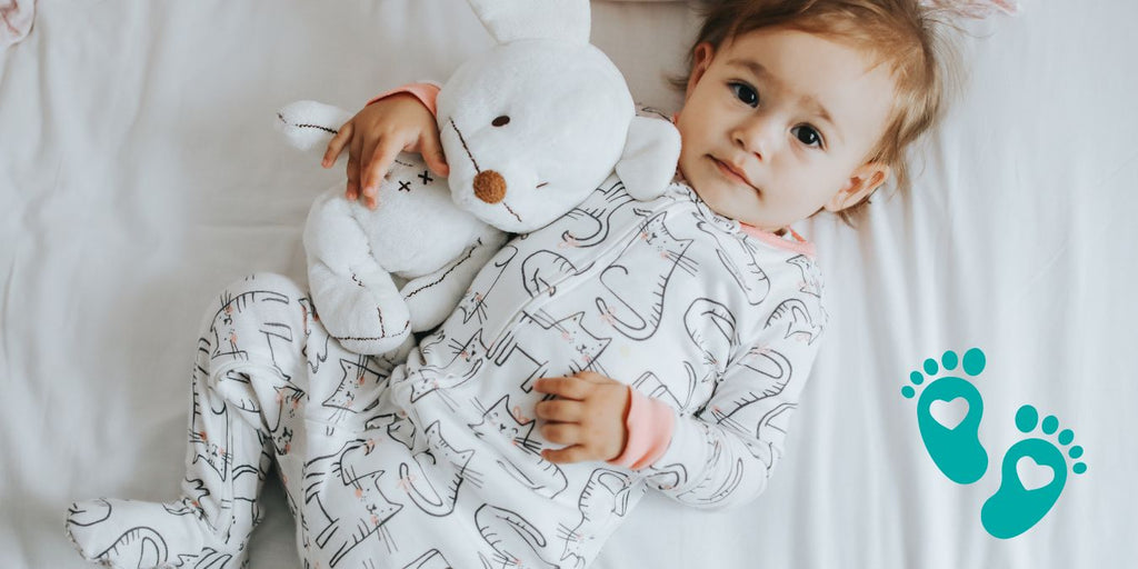 Toddler lying on a bed in a onesie with a stuffed animal, promoting Grookz baby shoes 