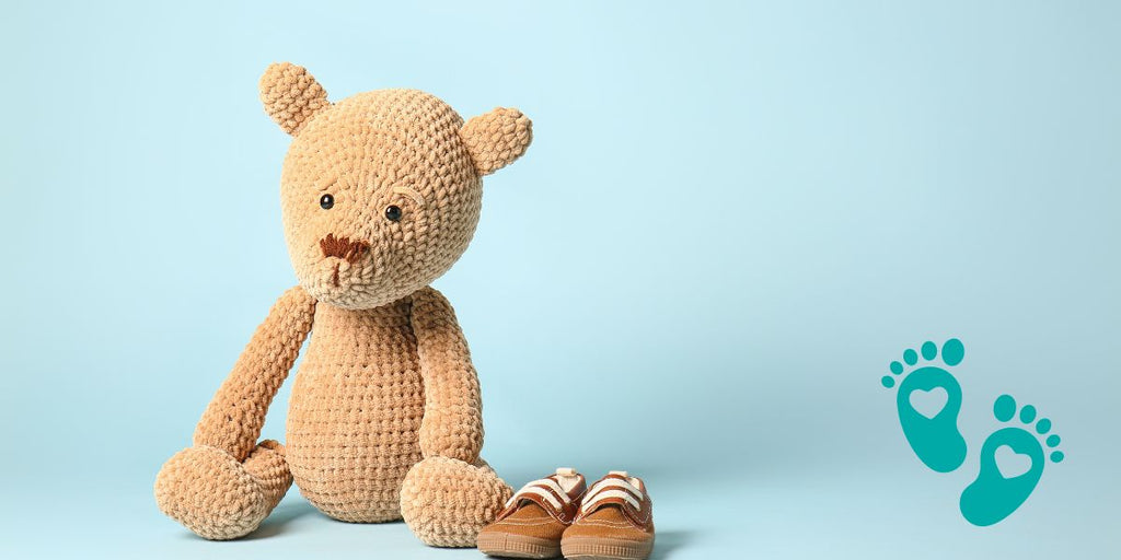 Knitted teddy bear sitting next to a pair of baby shoes on a light blue background, offering a guide to the trendiest baby shower gifts with Grookz Shoes