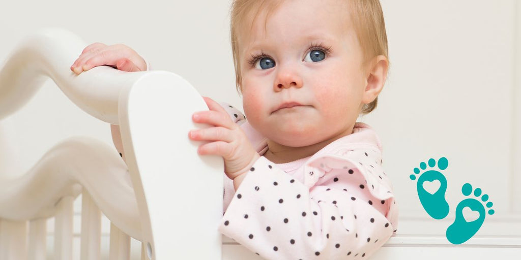 Curious 9-month-old baby girl standing in a crib, highlighting Grookz baby shoes with teal footprints logo.