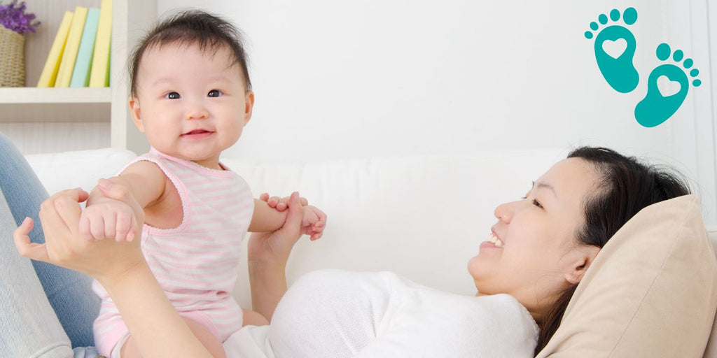 Smiling baby girl held by mother while lying on a couch, showcasing Grookz baby shoes with teal footprints logo.