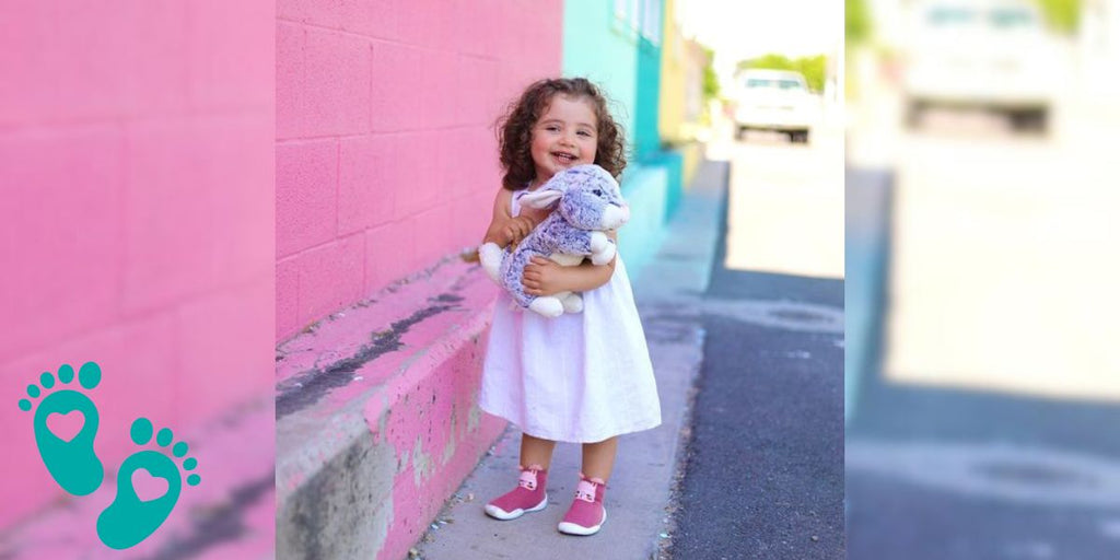 Little girl in a white dress holding a stuffed animal, standing by a colorful wall, exploring why baby shoes are so hard to put on with Grookz Shoes